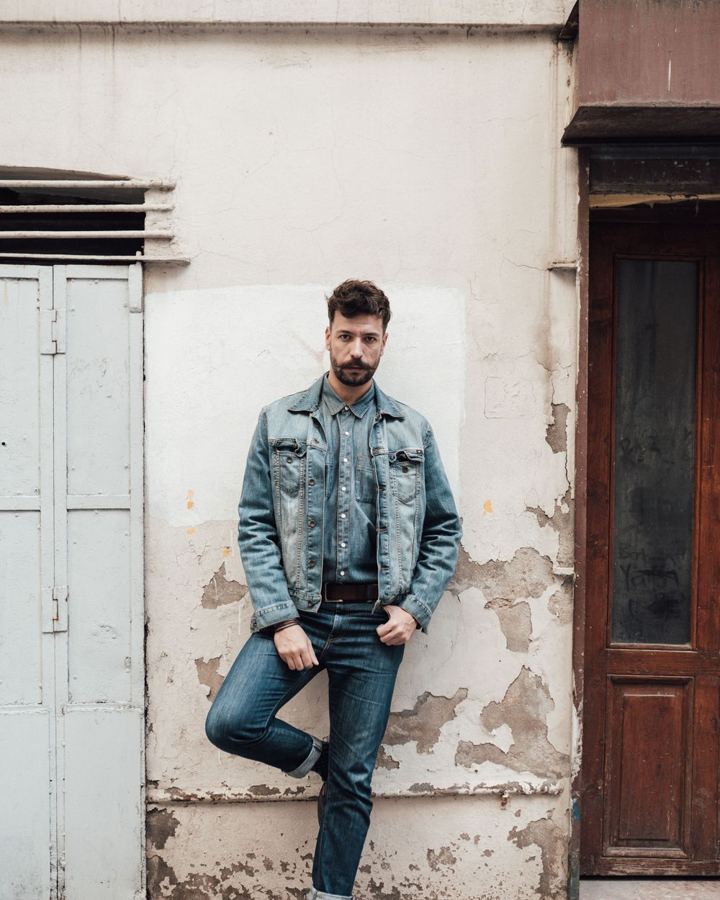 Man in a light denim jacket and dark jeans leaning on a wall